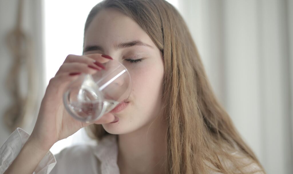 A young woman with brown hair drinks water from a clear glass indoors, eyes closed.