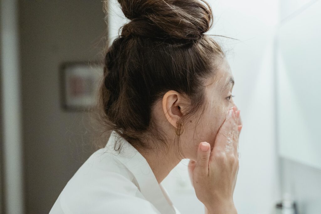 A young woman in a bathrobe follows her skincare routine, washing her face in a bathroom.